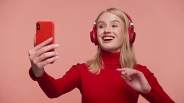 Kırmızı kulaklık takan gülümseyen genç bir kadın bir video görüşmesinde şeftali arka planına karşı akıllı telefon kullanıyor..