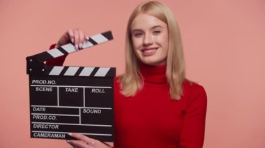 Kırmızı balıkçı yaka kazaklı genç bir kadın elinde bir klape tutarken gülümsüyor, film yapımcılığını ya da video yapımcılığını simgeliyor..