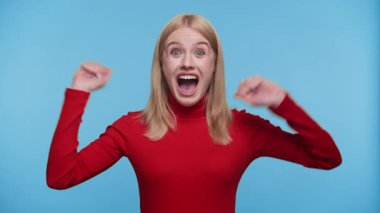 Kırmızı kazaklı heyecanlı genç kadın sevinçle tezahürat yapıyor, mutluluğu ifade ediyor ve mavi bir arka planda kutlama yapıyor..