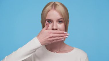 Tarafsız yüzlü sarışın bir kadın eliyle ağzını kapatıyor. Mavi bir arka planda sessizlik ya da gizlilik sinyali veriyor..