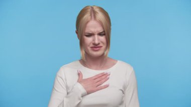 Beyaz gömlekli genç bir kadın göğsünde gözle görülür bir ağrı var. Rahatsızlık belirtileri gösteriyor. Ya da mavi arka planda muhtemel kalp sorunu..