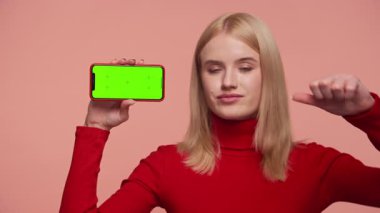 Etkilenmemiş genç bir kadın yeşil ekranlı akıllı telefonu tutuyor ve hayal kırıklığını ya da uygulamayı onaylamadığını belirterek baş parmağını kaldırıyor..