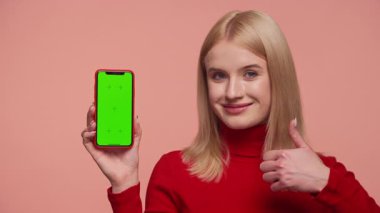 Kırmızı kazaklı, gülümseyen kadın yeşil ekran maketli akıllı telefonu tutuyor ve pembe arka plana başparmak işareti veriyor..