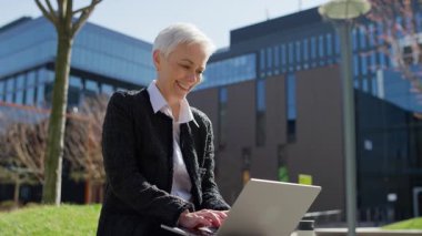 Kendinden emin yaşlı bir kadın, güneşli bir günde gülümseyen, modern bir ofis binasının dışında dizüstü bilgisayarla uzaktan çalışmaktan zevk alıyor..