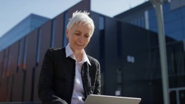 Dışarıda çalışan neşeli yaşlı iş kadını, modern bir ofisin önünde dizüstü bilgisayar kullanırken heyecan içinde yumruğunu kaldırıyor..
