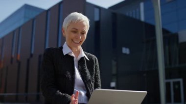 Neşeli yaşlı bir kadın, güneşli bir günde modern bir ofis binasının önünde bilgisayarında uzaktan bir video görüşmesi yapıyor..