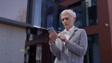 Endişeli yaşlı kadın, modern bir ofis binasının önünde dururken akıllı telefonundan üzücü bir mesaj okuyor..