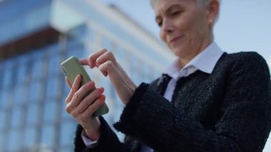 Arka planda modern cam binalarla dışarıda dikilirken akıllı telefon ekranını dinleyen yaşlı kadının yakın çekimi..