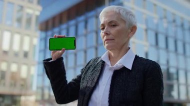 Yaşlı bir kadın modern cam ofis binasının önünde yeşil ekranlı ve nötr yüzlü akıllı telefon gösteriyor..