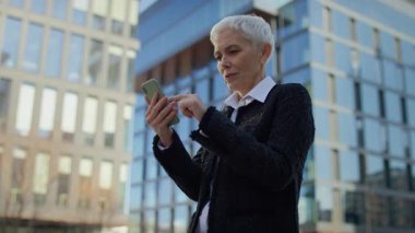 Yaşlı kadın, güneşli bir günde modern cam ofis binalarının önünde akıllı telefon kullanırken gülümsüyor..