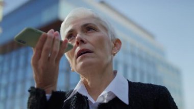 Camdan ofis binalarının yakınlarındaki bir telefon konuşması sırasında akıllı telefonu hoparlörde tutan yaşlı iş kadını..