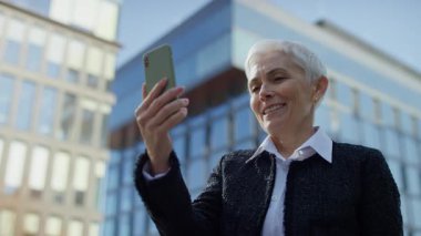 Akıllı telefonuyla konuşan ve modern cam ofis binalarının önünde dikilen olgun bir kadın..