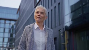 Olgun bir iş kadını işe giderken güvenli bir şekilde modern cam binaların yanında yürüyor, profesyonel giysiler içinde..