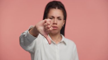 Memnun olmayan genç bir kadın ciddi bir ifadeyle baş parmak hareketi yapıyor, pembe bir arka planın önünde duruyor..