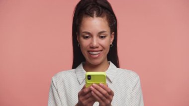 Heyecanlı genç bir kadın, pembe bir arka planda dururken yüzünü tutarak, sürpriz ve neşeyle akıllı telefonuna bakar..