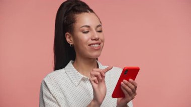 Diş teli takan genç bir kadın, elinde pembe bir arka planda kırmızı bir akıllı telefon tutarak online alışveriş tercihini değerlendiriyor..
