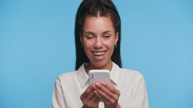 Heyecanlı genç kadın akıllı telefonunu kaydırırken parlak bir şekilde gülümsüyor, mavi arka planda izole edilmiş online içerik veya haberlere mutlu bir tepki gösteriyor..