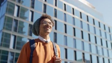 Sırt çantası ve beresiyle gülümseyen genç adam cam binalarla dolu bir şehirde yürüyor, güneşli bir şehir gününün tadını çıkarıyor..