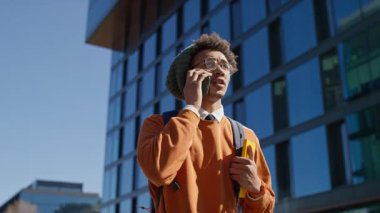 Sırt çantası ve not defteri olan odaklanmış genç adam güneşli bir günde modern cam binaların önünden geçerken akıllı telefondan konuşuyor..