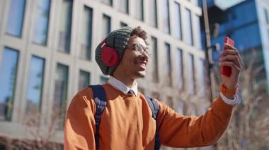 Kulaklık ve sırt çantası takan gülümseyen genç adam, modern şehir bölgesinde yürürken akıllı telefon kullanarak bir video görüşmesinde konuşuyor..