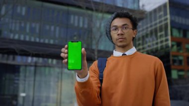 Ciddi bir genç adam kameraya doğru yeşil ekranlı akıllı bir telefon sunar, modern cam binaların önünde durur..