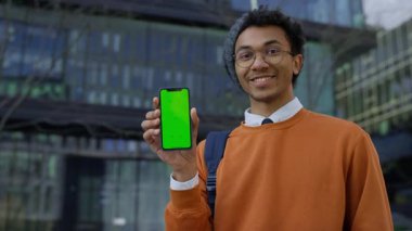 Neşeli genç adam yeşil ekranlı akıllı bir telefonu modern şehir ortamının dışında dururken onaylamak için başını sallıyor..