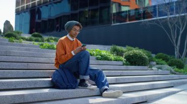 Modayı takip eden genç adam şehir basamaklarında oturuyor, günlük kıyafetler giyiyor ve beresini takıyor, modern bir şehir ortamında akıllı telefonunu karıştırıyor..
