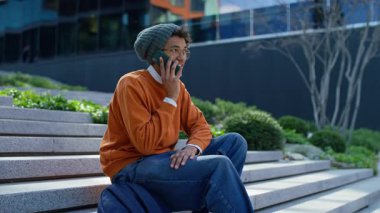 Modern taş basamaklarda oturan, yeşillik ve binalarla dolu bir şehir ortamında telefon görüşmesi yapan sıradan bir genç adam..