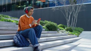 Basamaklarda oturan, kulaklık takan ve bereli genç bir adam modern bir şehir ortamında akıllı telefonuyla düşünceli bir şekilde kayıyor..