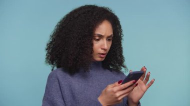 Kıvırcık saçlı genç bir kadın, üzgün görünüyor. Akıllı telefonuyla üzücü haberler okuyor ya da mavi arka planda mesajlar bırakıyor..