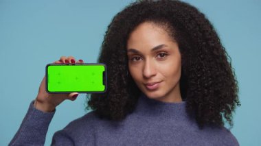 Kendine güveni tam genç bir kadın, yeşil ekran ekranlı akıllı telefonu açık mavi arkaplana karşı uygulama veya içerik modellemesi için hazır tutuyor..
