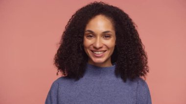 Kıvırcık saçlı, mutlu bir genç kadının portresi. Yumuşak pembe bir arka planda sıradan bir süveterle dururken sıcak bir şekilde gülümsüyor..