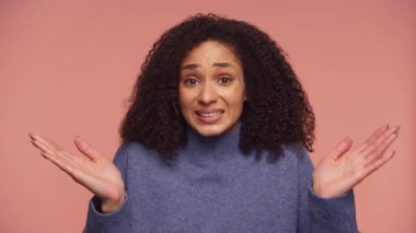 Mavi kazaklı genç bir kadın pembe bir arka plana karşı omuz silkerek kafa karışıklığı ya da belirsizlik gösteriyor..