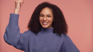 Kıvırcık saçlı neşeli genç kadın heyecan ve zafer ifade ediyor. Parlak bir şekilde gülümsüyor ve pembe bir arka plana karşı yumruğunu kaldırıyor..