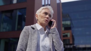 İş kıyafetleri içindeki kendinden emin yaşlı kadın modern ofis binalarının dışında akıllı telefondan konuşuyor ve ciddi bir sohbete girişiyor..