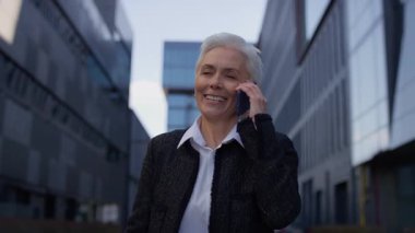 Beyaz saçlı neşeli yaşlı bir kadın modern bir şehir manzarasında yürüyor, bir telefon konuşması sırasında gülümsüyor..