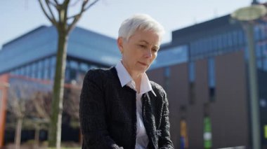 Düşünceli yaşlı kadın bilgisayarını modern bir şehir ortamında kullanırken düşünmek için duruyor, doğal ışıkta uzaktan çalışmanın keyfini çıkarıyor..