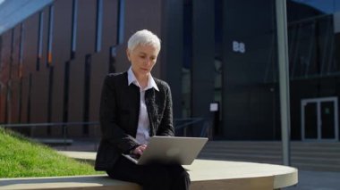 Yaşlı bir kadın, modern bir şehir ortamında dizüstü bilgisayarla çalışırken sinirli bir ifade sergiliyor. Teknolojik bir sorunla karşı karşıya..