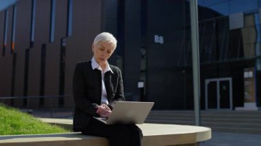 Yaşlı kadın modern bir şehir bölgesinde oturuyor, dizüstü bilgisayarındaki video görüşmesi sırasında parmağını kaldırıyor ve konuşuyor..