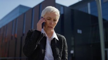 Olgun bir iş kadını, açık hava ortamında dizüstü bilgisayarla otururken akıllı telefonuyla güvenle konuşur..