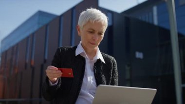 Açık havada kredi kartı ve laptop kullanan kendinden emin yaşlı bir kadın, muhtemelen online alışveriş ya da banka işlemi yapıyor..