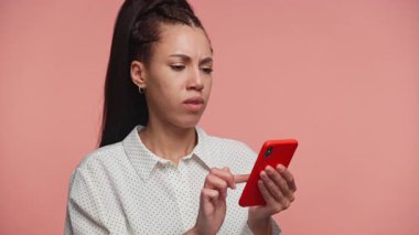 Sinirli genç bir kadın, telefonunun ekranına bakarken hayal kırıklığı yaratan veya negatif içeriğe tepki veriyor, pembe arka planda izole edilmiş..