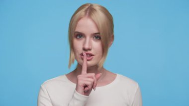 Beyaz gömlekli genç bir kadın, mavi arka plana karşı sessiz kalmak için parmaklarını dudaklarına dayayarak sessizliği işaret ediyor..