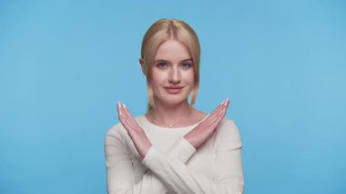Beyaz elbiseli genç kadın kollarını önünde kavuşturarak poz veriyor, hayır işareti veriyor, inkar ediyor veya mavi arka plana karşı reddediyor..