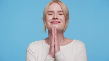 Minnettar, genç bir kadın, gözleri kapalı, rahatlama ve dua jesti ile gülümsüyor, mavi arka planda umut ya da şükran duygularını ifade ediyor..