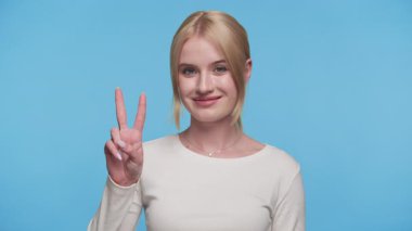 Gülümseyen genç bir kadın parmaklarıyla barış ya da zafer işareti yapıyor, mavi bir arkaplana yaslanmış beyaz bir bluzla..