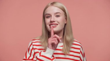 Çizgili tişörtlü sarışın kadın parmaklarını dudaklarına uzatarak gülümsüyor. Pembe arka plana karşı sessizlik ya da gizlilik işareti yapıyor..