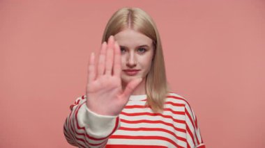 Çizgili tişörtlü sarışın kadın dur işaretinde el ele tutuşuyor. Pembe arka plana bakıp uyarı ya da reddetme işareti veriyor..