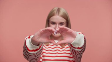 Çizgili tişörtlü sarışın kadın, pembe arka planda aşkı ya da sevgiyi sembolize eden elleriyle bir kalp şekli oluşturur..