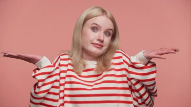 Çizgili gömlekli sarışın kadın ellerini kaldırmış ve şaşkın bir ifade takınarak pembe arka plana karşı kafa karışıklığı ya da umursamazlık gösteriyor..
